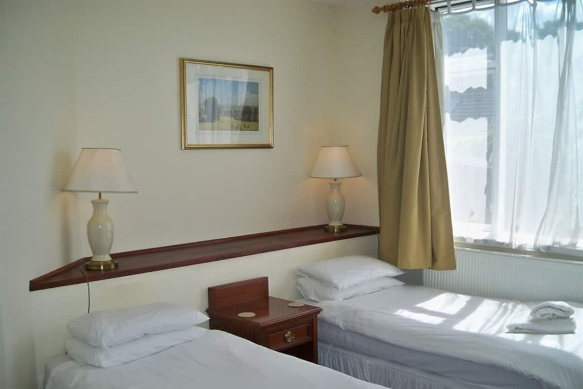 Habitación de hotel con dos camas individuales, una mesita de noche de madera, un cuadro en la pared y una ventana con cortinas.