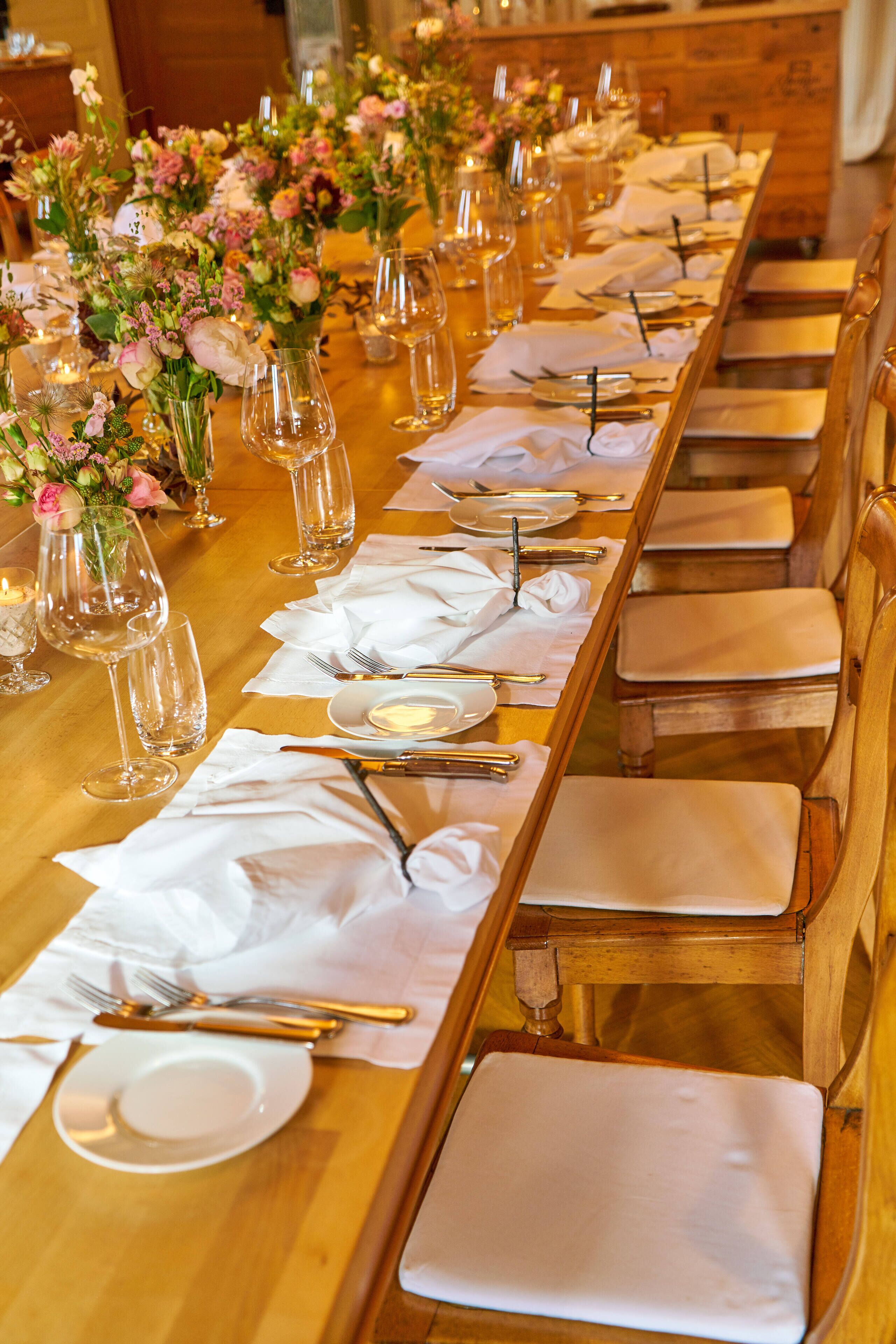 A long dining table set with white napkins, plates, cutlery, and wine glasses, flanked by chairs with cushioned seats.