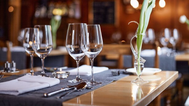 Un ambiente de restaurante con una mesa dispuesta para comer, destacando copas de vino, un florero con una flor y una vela.