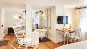 Una habitación con piso de madera, paredes blancas, una bañera blanca, un escritorio de madera con una silla, un televisor y un espejo.