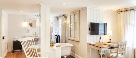 A room with a wooden floor, white walls, a white bathtub, a wooden desk with a chair, a television, and a mirror.