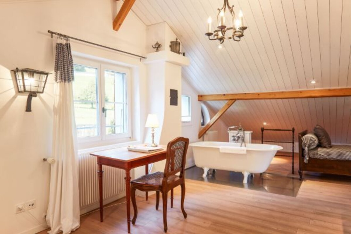 Una habitación con suelo de madera, una bañera blanca, una silla de madera, una mesa con lámpara y una ventana con cortinas.
