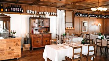 A restaurant with wooden interiors, multiple dining tables set with white tablecloths and wine glasses, and a variety of bottles on shelves.