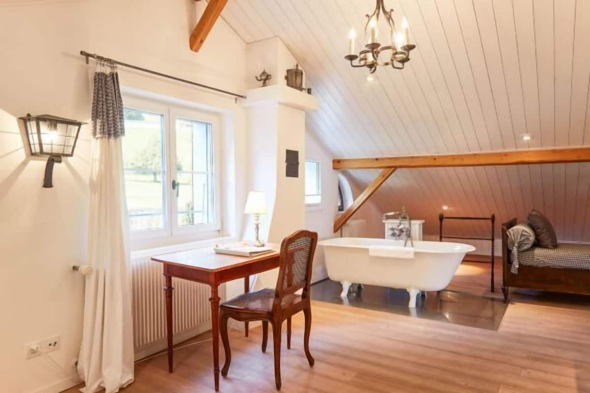 Ein Zimmer mit Holzboden, einer weißen Badewanne, einem Holzstuhl, einem Tisch mit Lampe und einem Fenster mit Vorhängen.