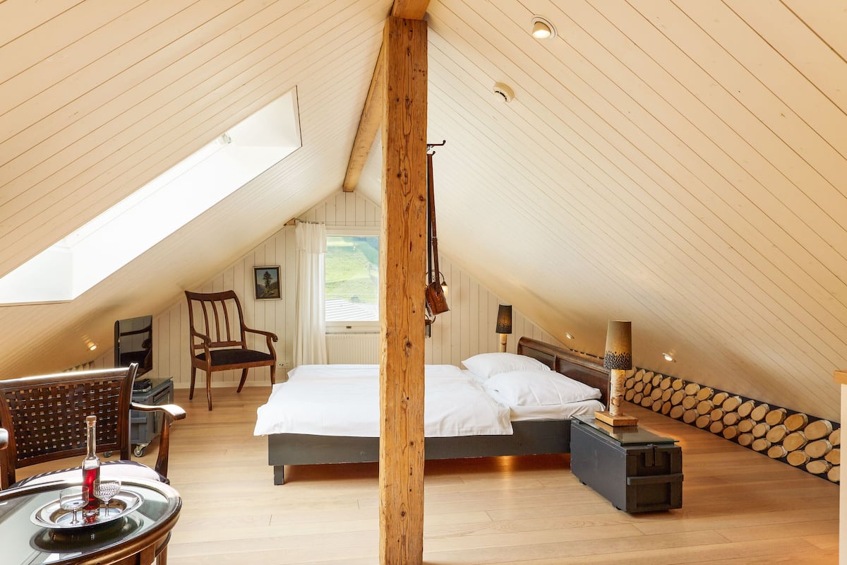 Ein Schlafzimmer mit geneigter Decke, Holzbalken, einem Bett mit weißen Leinen, einem Sessel, einem Tisch mit einer Flasche und einer Lampe.