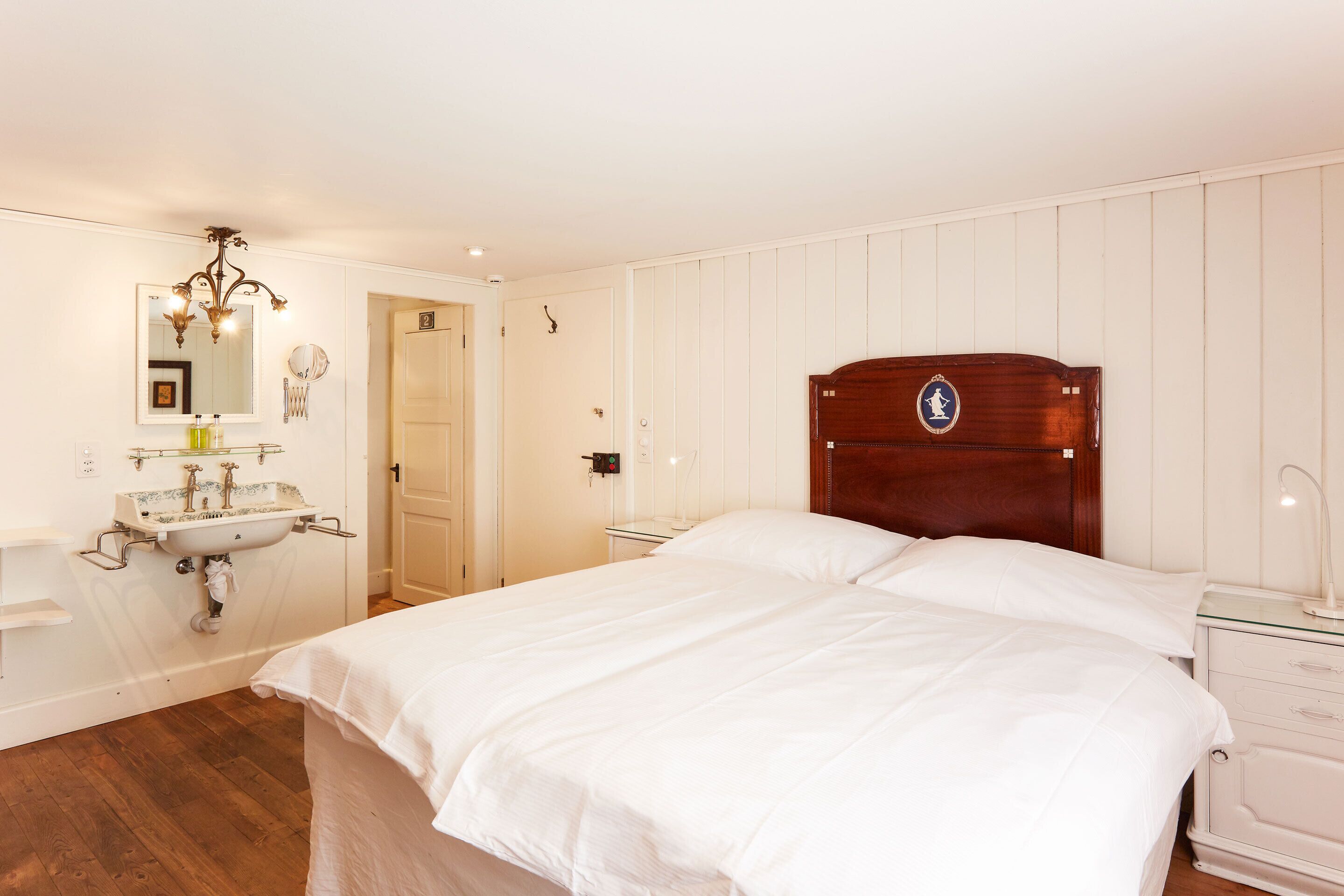 Een slaapkamer met een houten hoofdbord, een wit bed, een nachtkastje, een witte dressoir, een spiegel en een wastafel aan de muur.