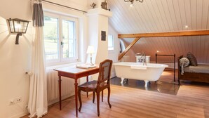 Una habitación con suelo de madera, una bañera blanca, una silla de madera, una mesa con una lámpara y una ventana con cortinas.