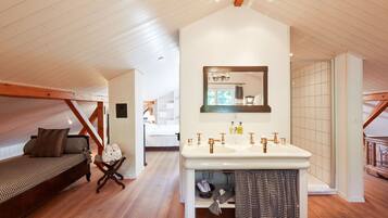 A bedroom with a wooden ceiling, a large window, a bed with a checkered bedspread, a bedside table with rolled towels, and a white sink with a mirror above it.