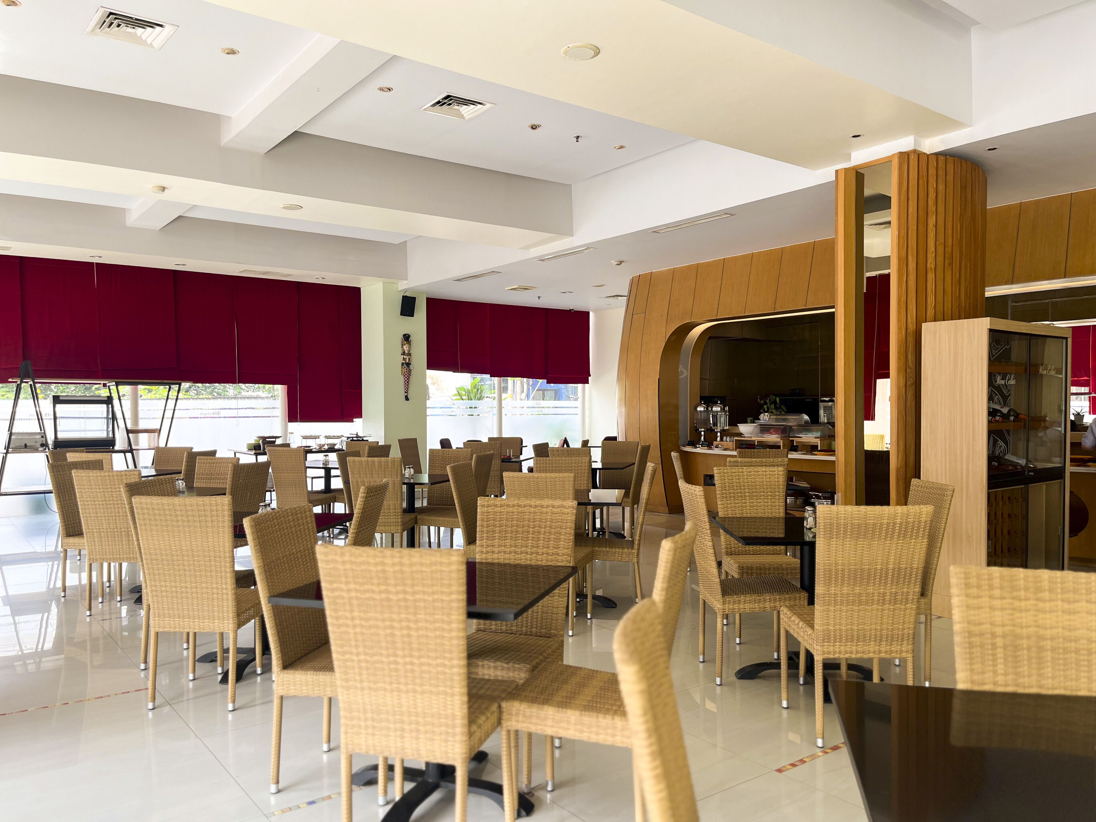 Un restaurant modern amb caires de vimet i taules, cortines vermelles, i una zona de cuina visible a través d'una arcada.