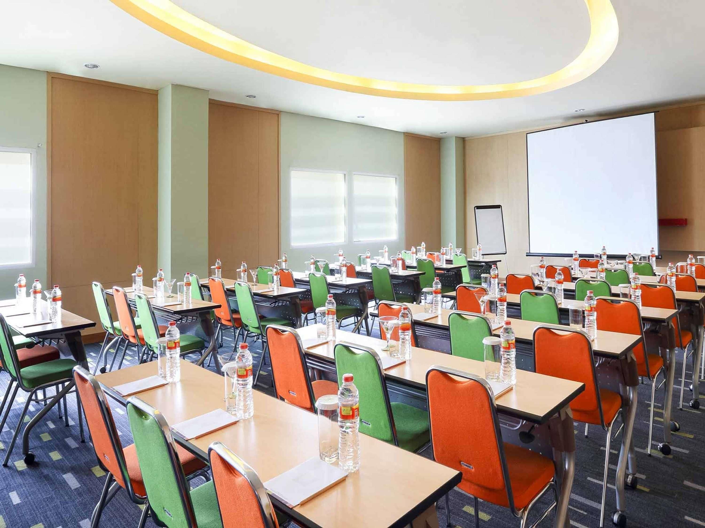 A conference room with multiple tables and chairs, equipped with water bottles and a projector screen.