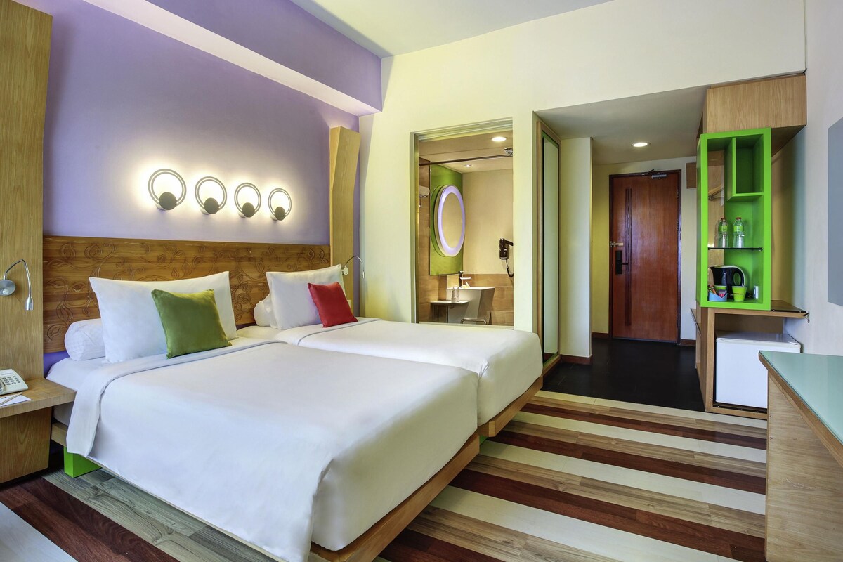 Une chambre d’hôtel avec deux lits, une tête de lit en bois, une table de chevet avec un téléphone, un miroir et un meuble de rangement vert.