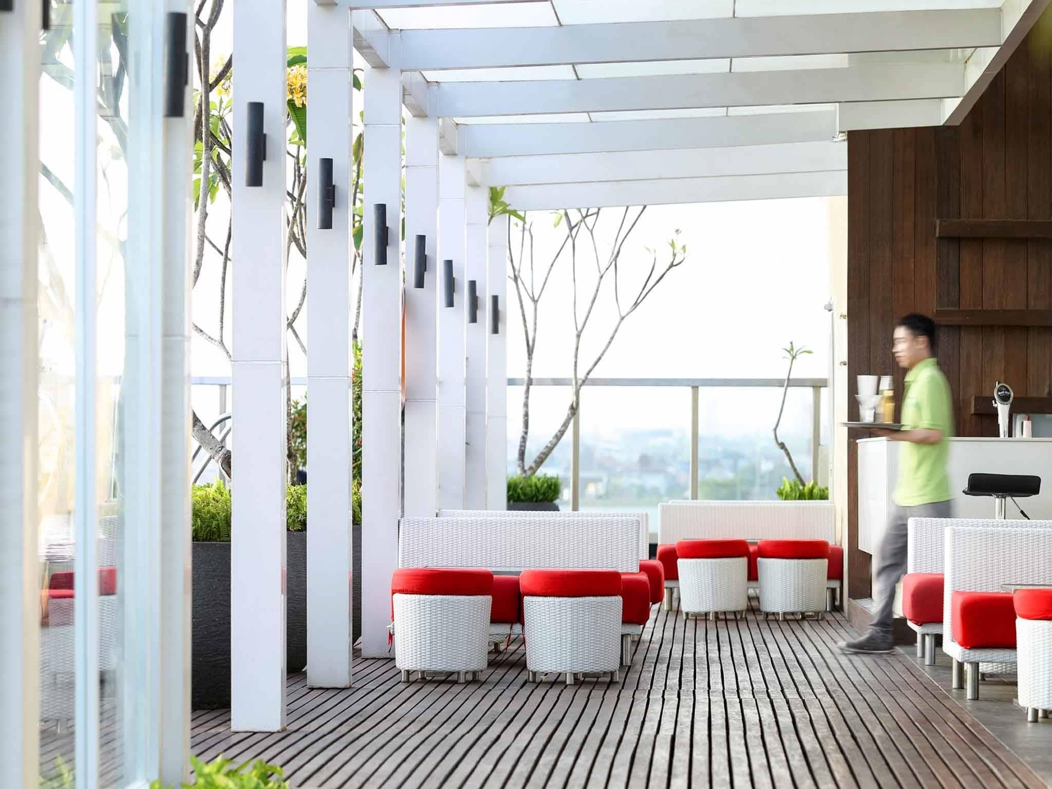 A modern outdoor seating area with white wicker furniture and red cushions, a wooden deck, and a view of the city.