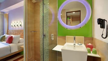 Une chambre d’hôtel moderne dotée d’une douche en verre, d’un lit avec des oreillers, d’une table de chevet, d’un miroir rond avec un éclairage intégré et d’un lavabo avec un distributeur de savon.