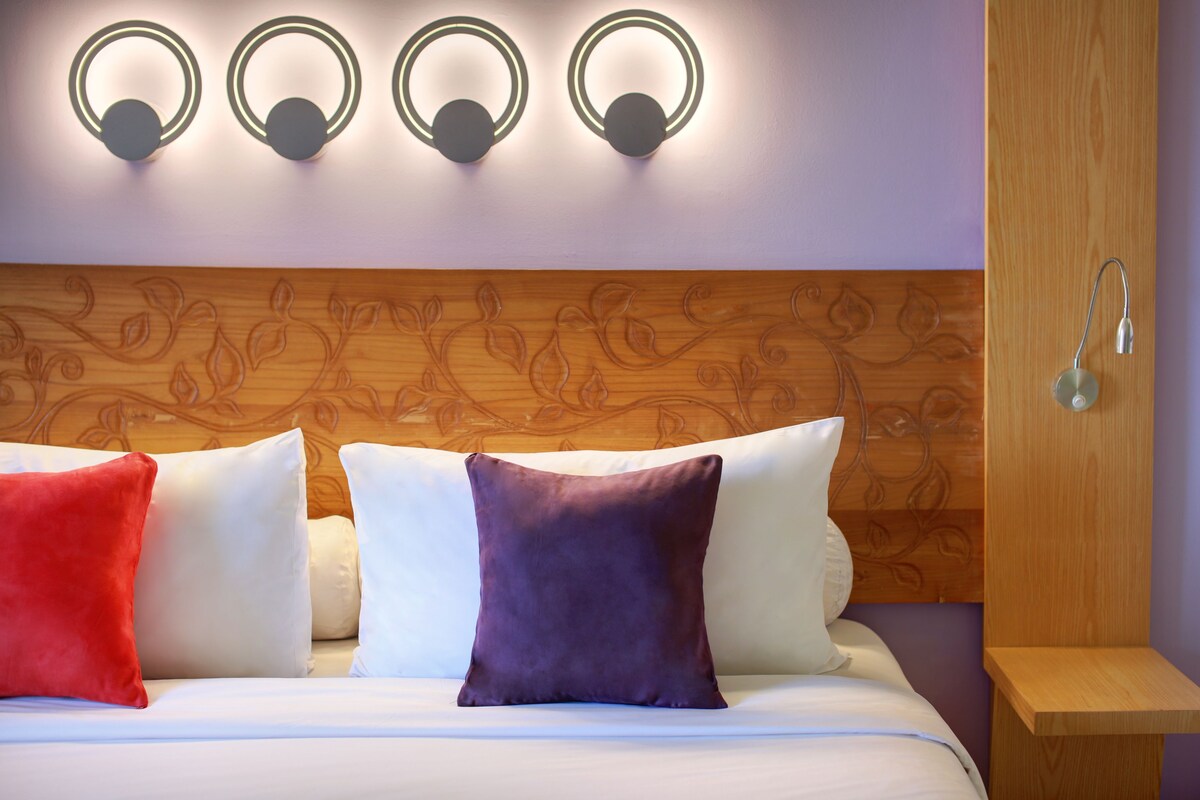 A neatly made bed with four pillows in different colors, a wooden headboard with carved designs, and wall-mounted lighting fixtures above the bed.