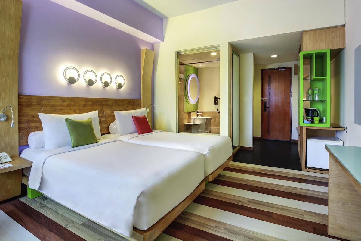 Quarto de hotel com duas camas, cabeceira de madeira, mesa de cabeceira com telefone, espelho e um móvel de armazenamento verde.