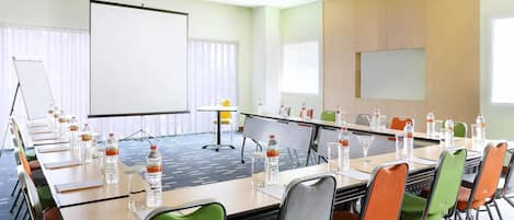 Un salón de conferencias con una larga mesa, sillas, una pantalla de proyector y botellas de agua.