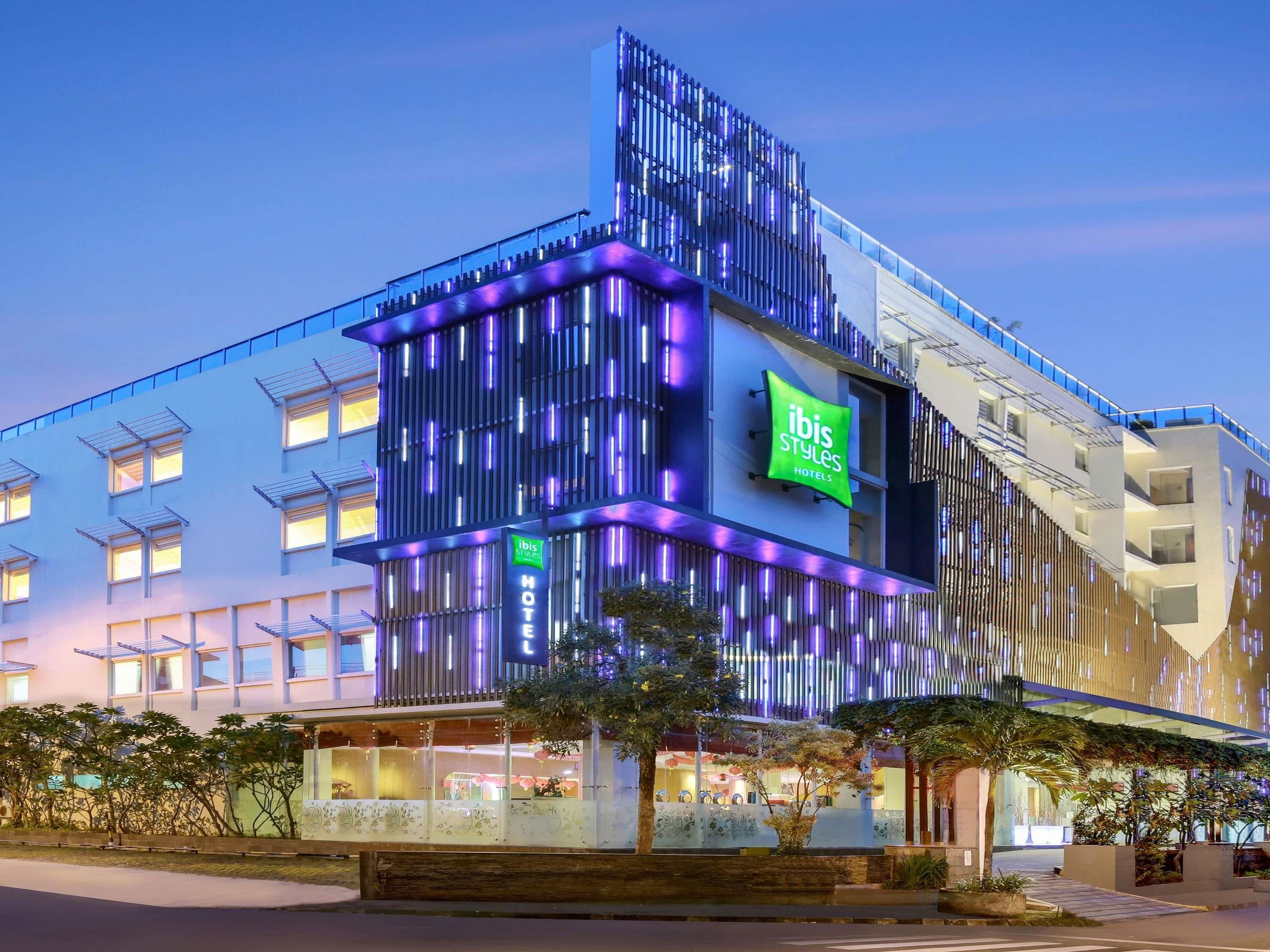A modern hotel building with a prominent 'ibis Styles Hotels' sign, multiple balconies, and a well-lit facade.