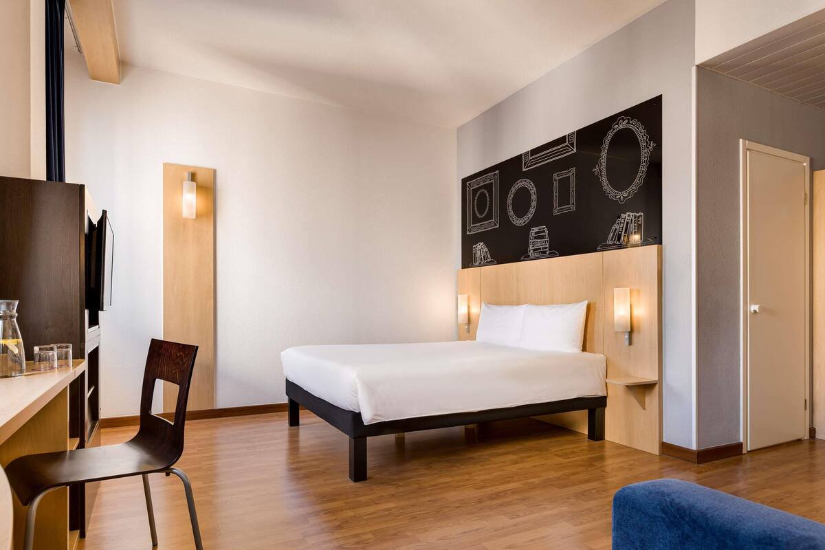 A hotel room with a bed, a desk with a chair, a coffee maker, and a wall with framed pictures.