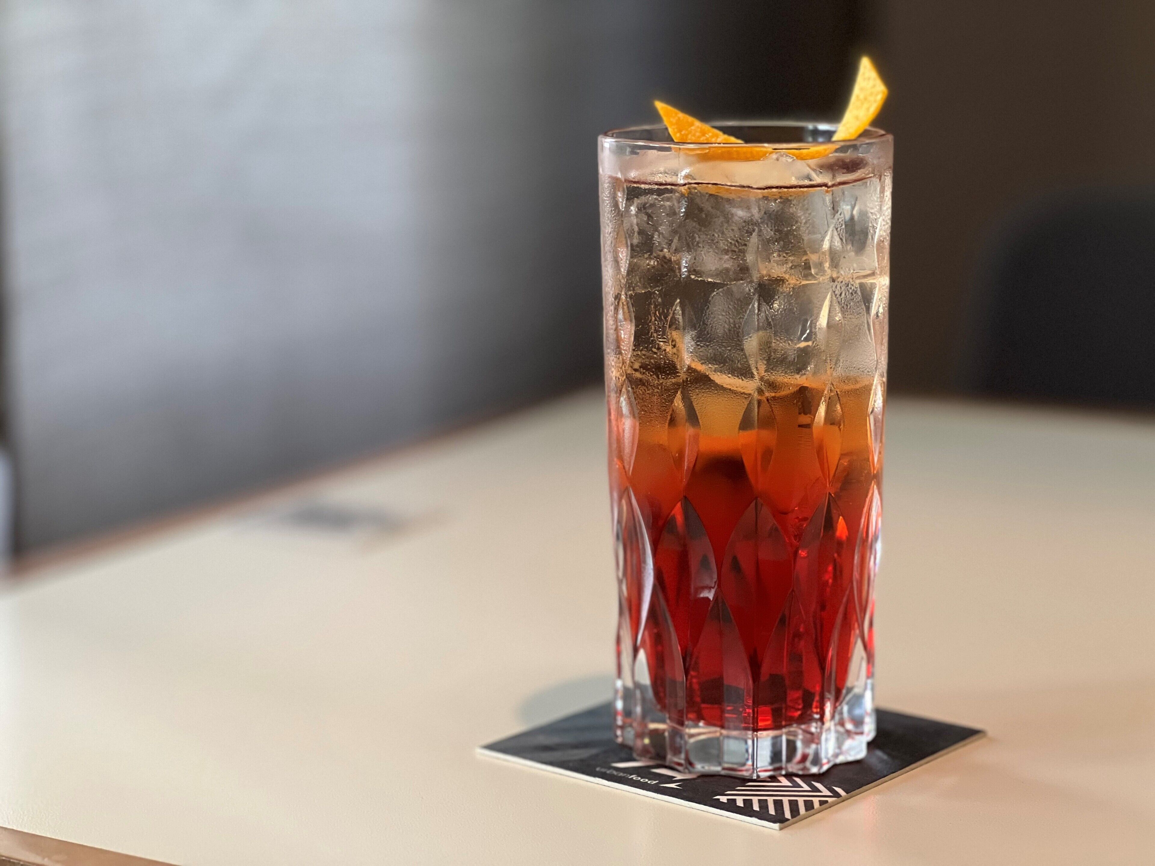Ein hoher, strukturierter Glasbecher gefüllt mit einem roten Getränk und Eiswürfeln, mit einer Orangenscheibe verziert, auf einem Untersetzer.