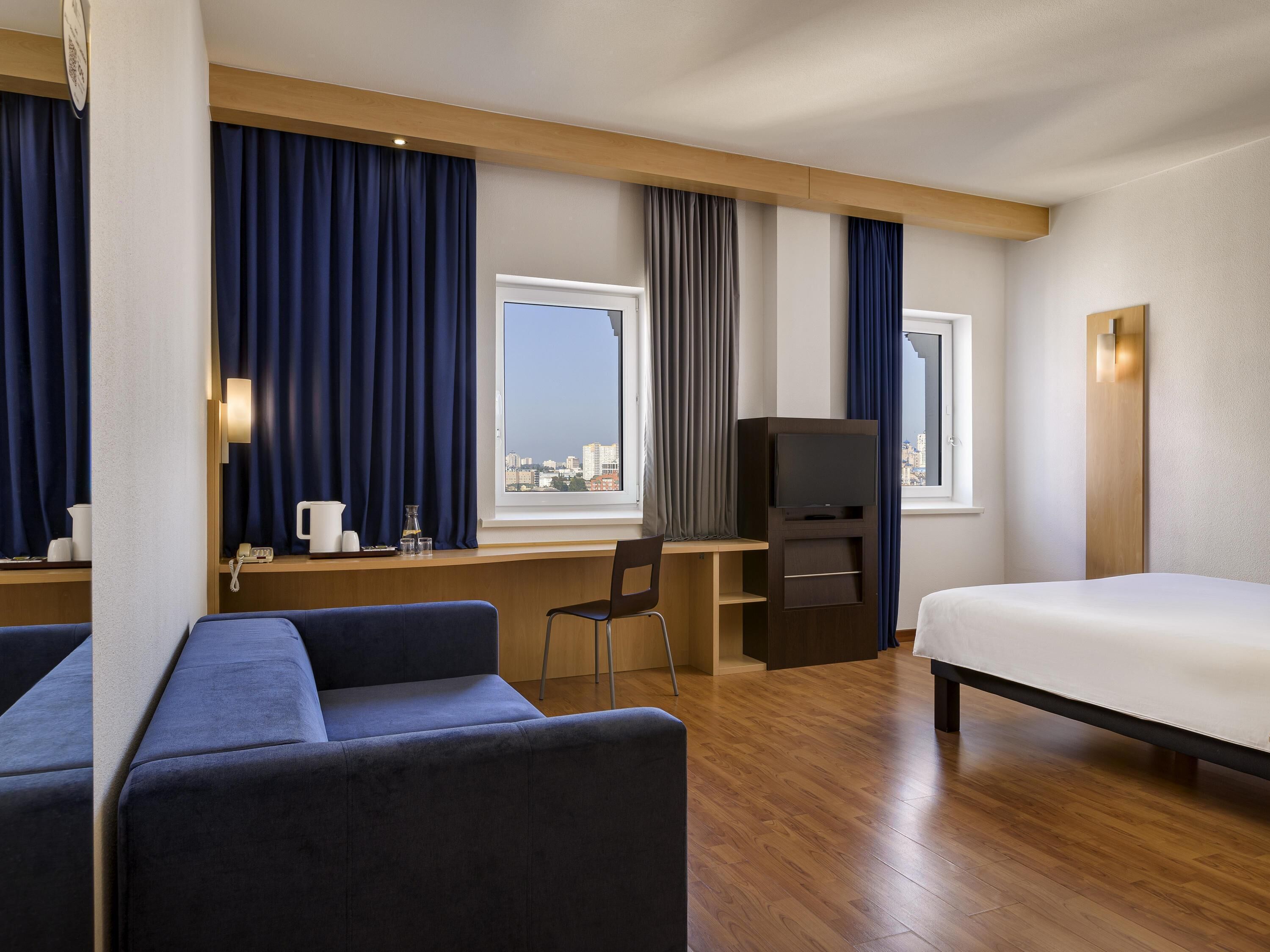 A hotel room with a bed, a desk, a chair, a TV, and a view of the city.