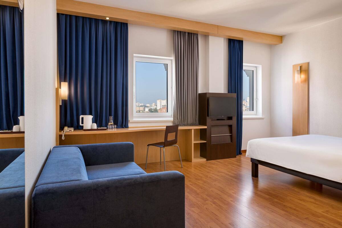 Ein Hotelzimmer mit Bett, Schreibtisch mit Computer, Stuhl, Fernseher und einem Fenster mit Vorhängen.