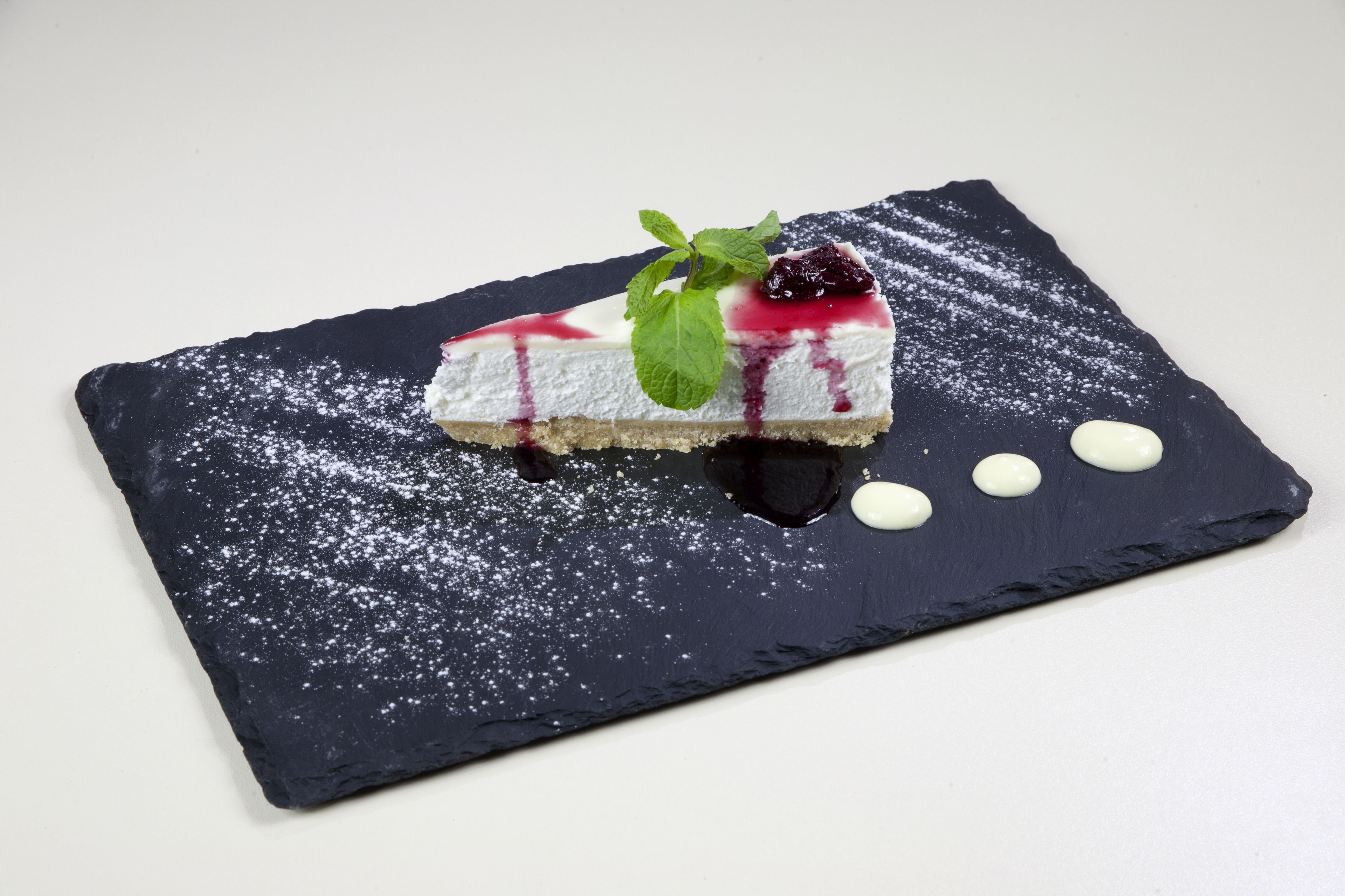 A slice of cheesecake on a slate plate with powdered sugar and a berry compote, garnished with a mint leaf.