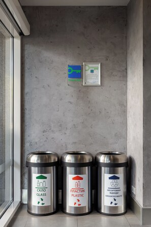 Three metal trash bins labeled for glass, plastic, and unsorted waste, placed against a concrete wall with a window and a framed document on the wall. - ibis Kyiv City Center (Kyiv)