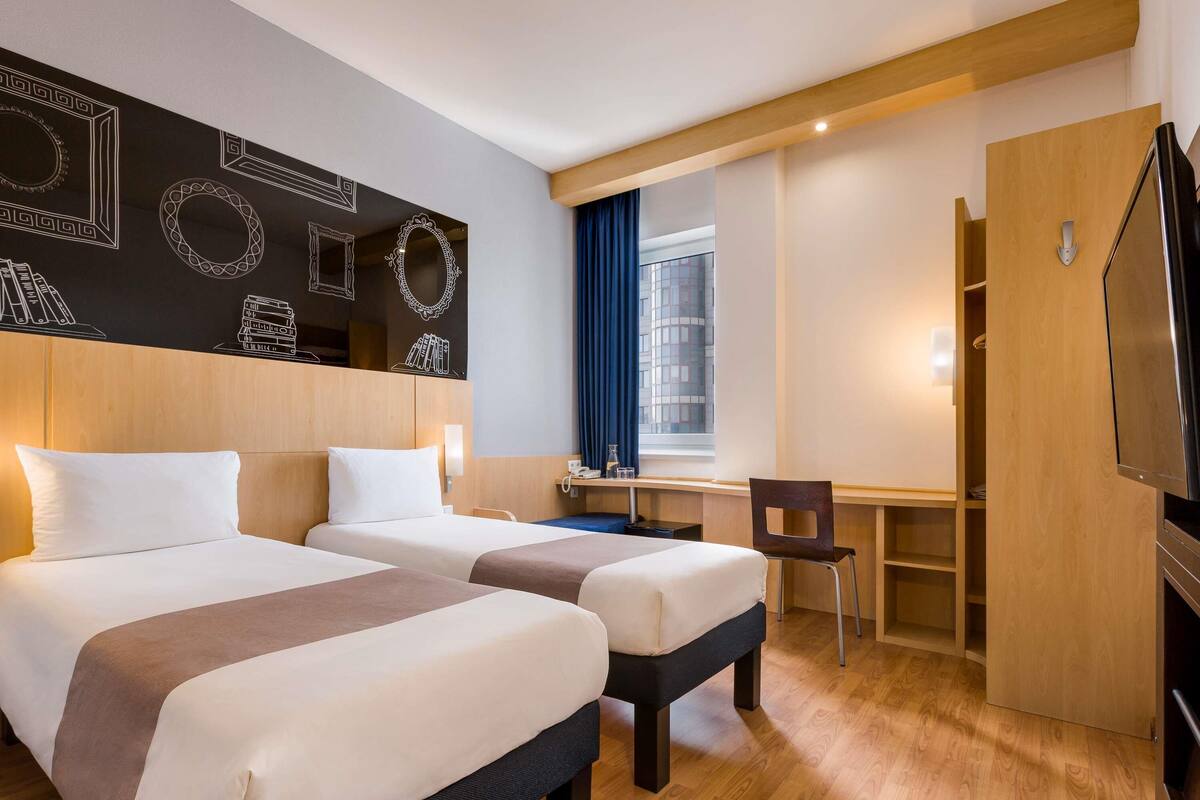 A hotel room with two beds, a desk, a chair, a TV, and a window with curtains.