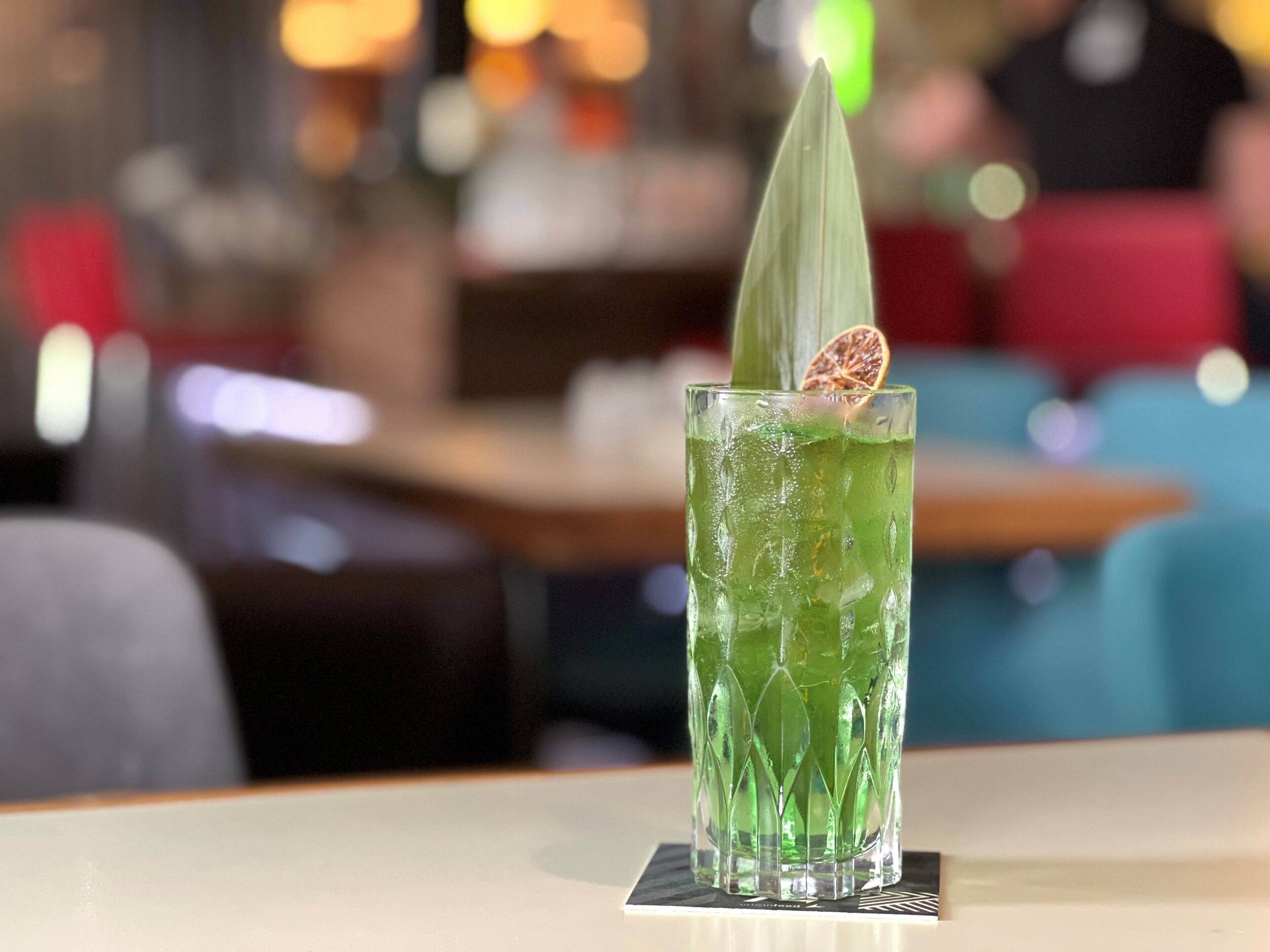 A tall glass with a green drink, garnished with a leaf and a slice of fruit, on a table in a restaurant.