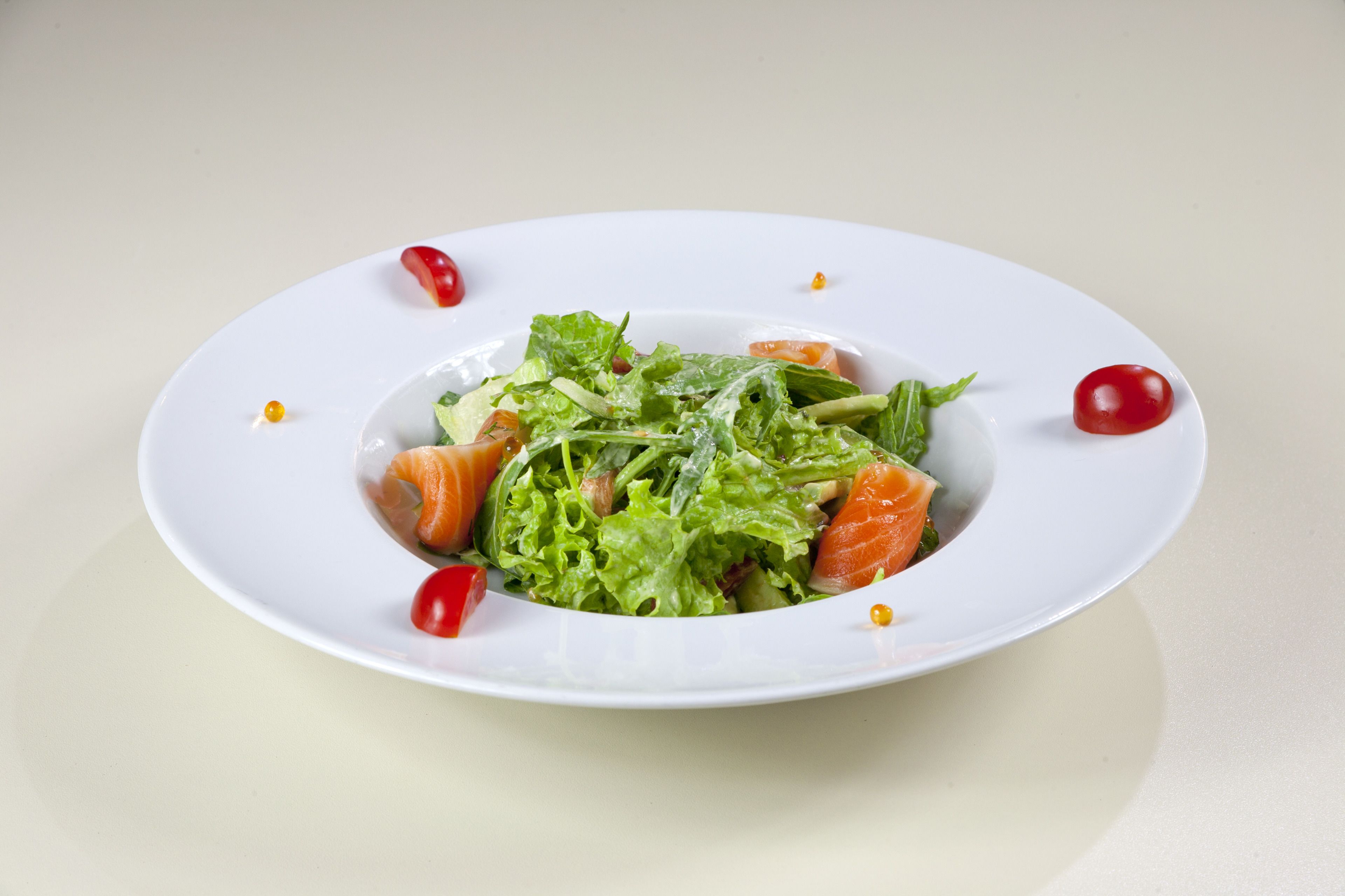 A white plate with a salad containing greens, tomatoes, and salmon, with a few cherry tomatoes and a lemon slice on the side.