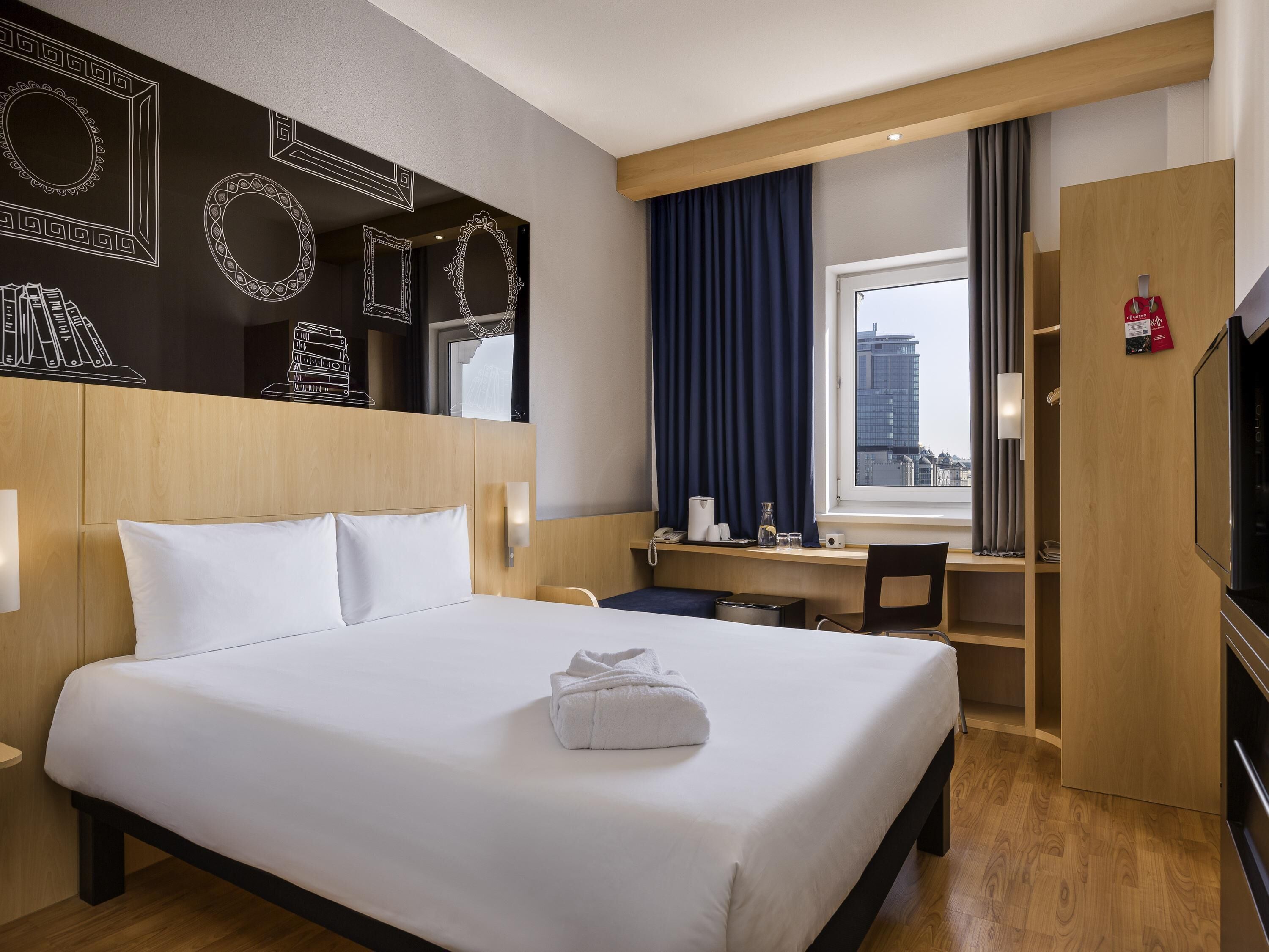 A hotel room with a bed, a desk, a chair, a TV, and a window with curtains.
