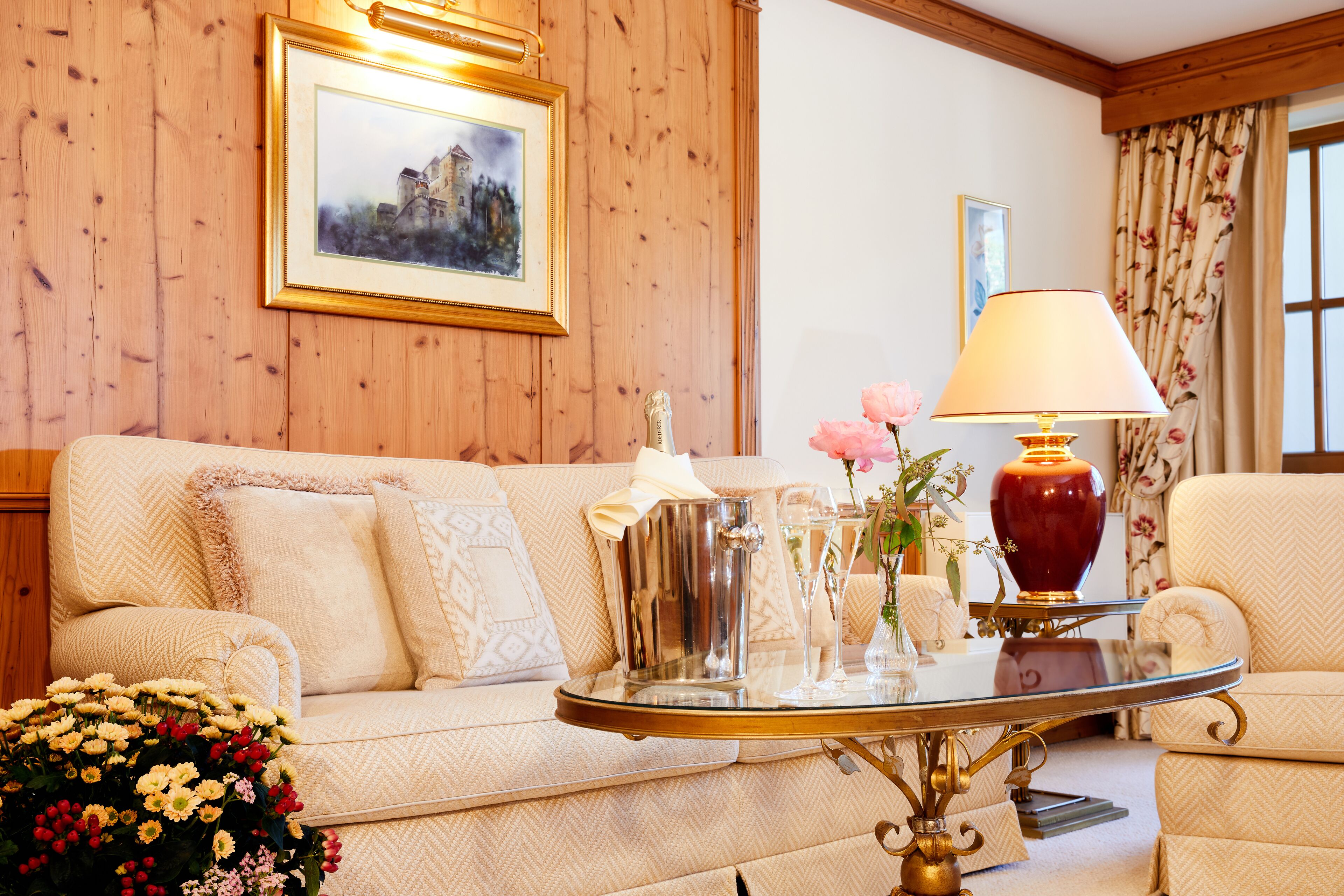 Una sala de estar acogedora con un revestimiento de madera, un sofá color crema, una mesa de centro de vidrio y un jarrón rojo con flores.
