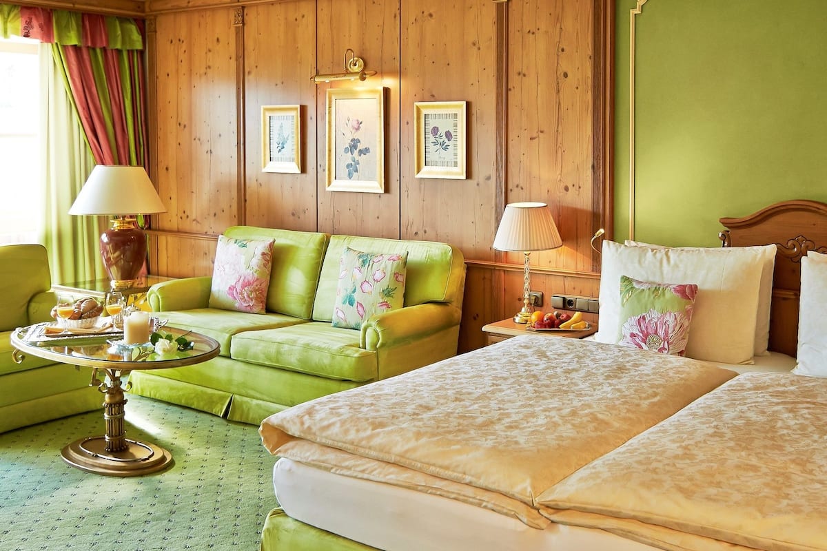 Een hotelkamer met een groene bank, een bed met bloemenbedrukking, een tafeltje met een ontbijttray, en bloemistische kunst op de muur.