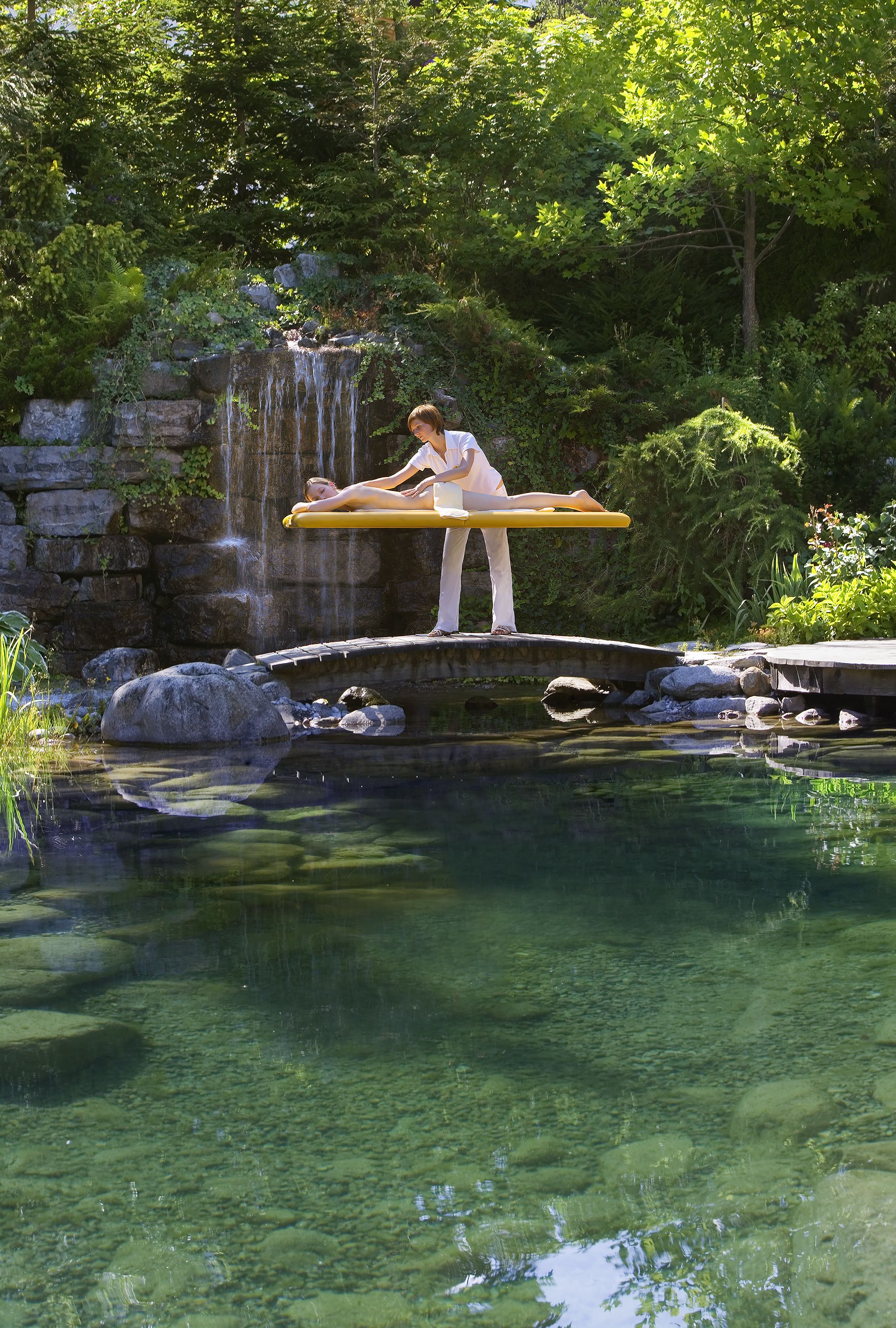 A person meditating on a wooden platform over a pond with a waterfall in the background.