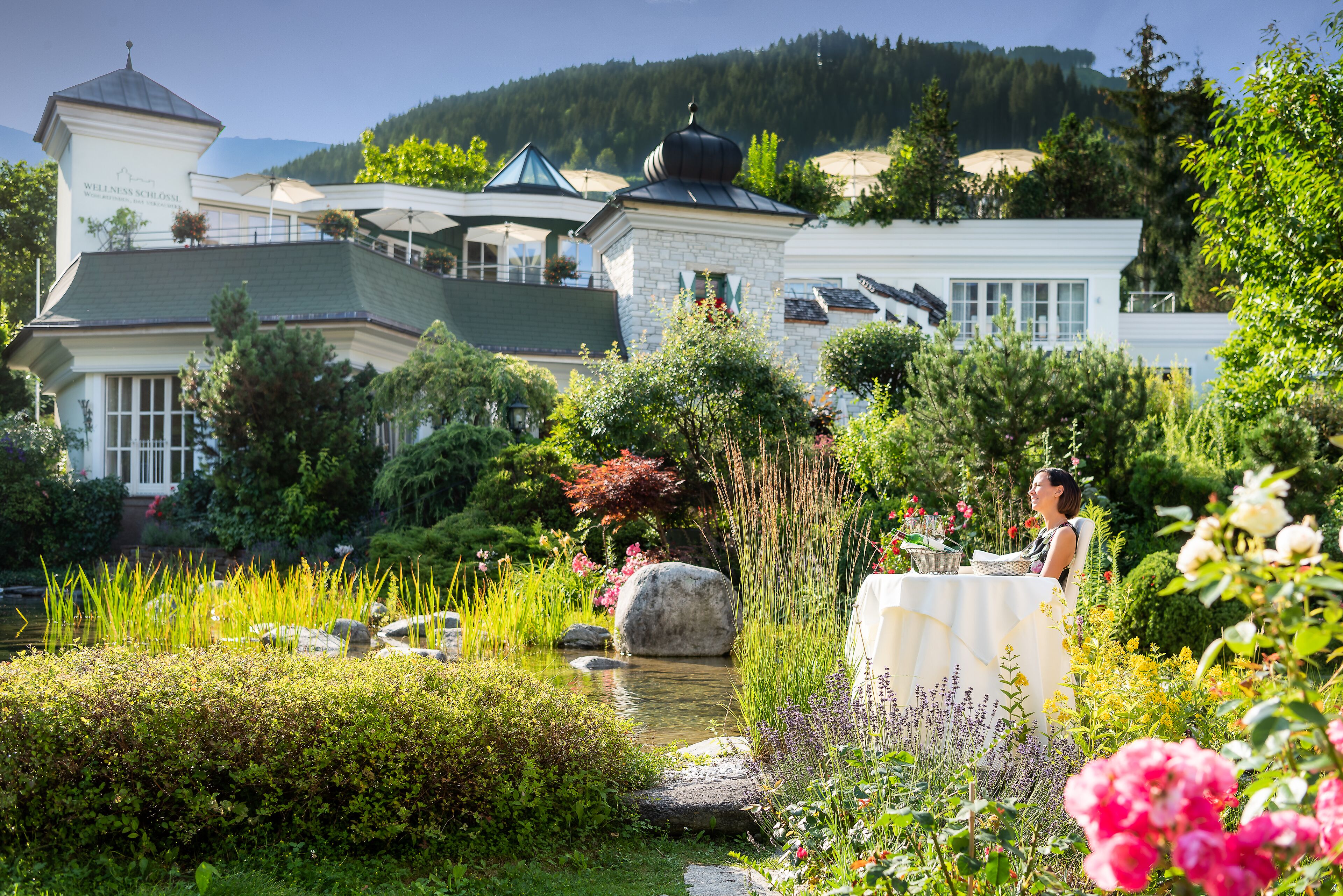A well-maintained garden with a pond, surrounded by lush greenery and flowers, and a building with a sign reading "Wellness Schloss."