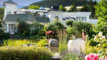 Un jardín bien cuidado con un estanque, rodeado de exuberante vegetación y flores, y un edificio con un letrero que dice 'Wellness Schloss'.