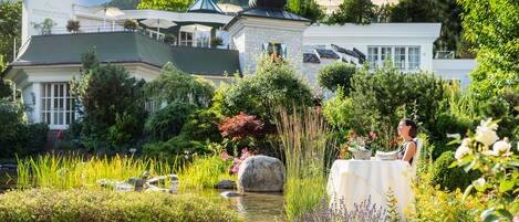 A well-maintained garden with a pond, surrounded by lush greenery and flowers, and a building with a sign reading "Wellness Schloss."