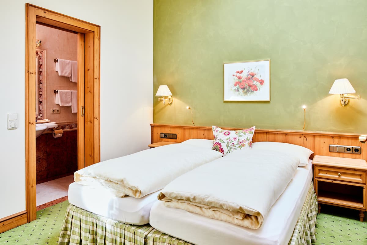 Een tweepersoonsbed met twee kussens, een houten nachtkastje, een wandlamp, een lijst met een bloemistisch afbeelding, en een deur naar een badkamer met handdoeken aan de muur.