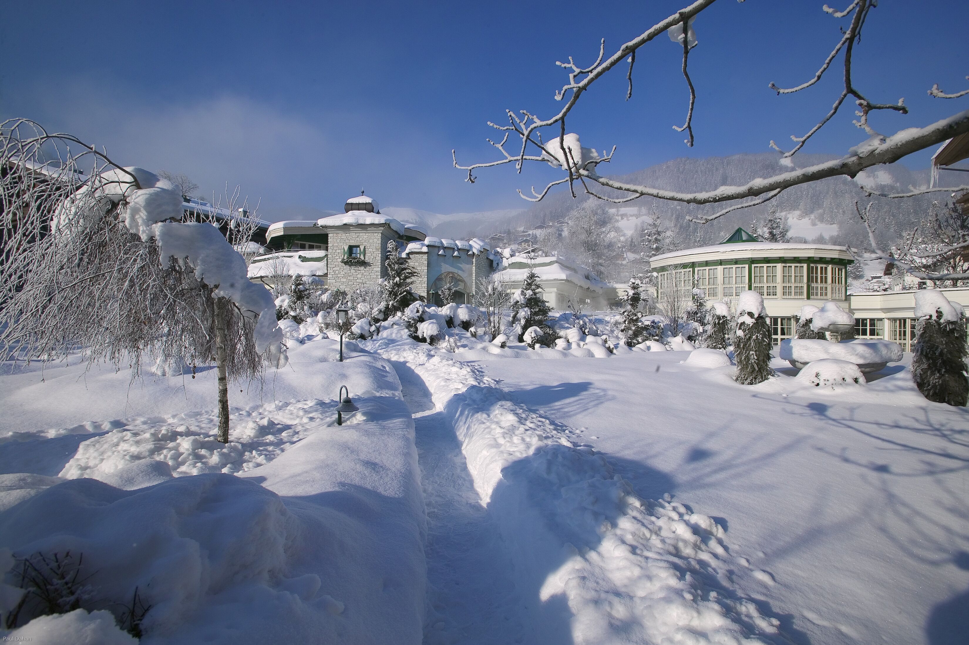 Schneebedeckte Landschaft mit Gebäuden, darunter eine Kirche und ein runder Bau, umgeben von Bäumen und Bergen im Hintergrund.