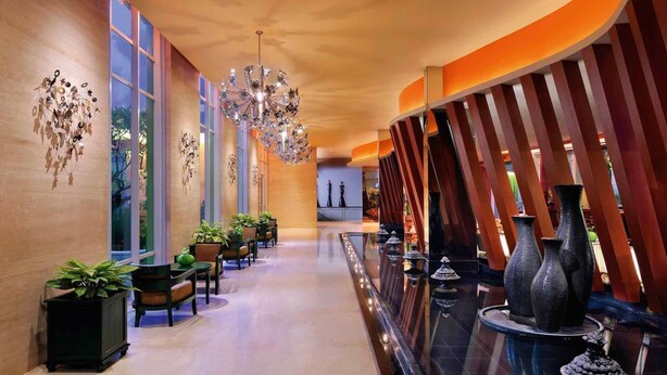 Un vestíbulo de hotel con un largo pasillo, ventanas amplias, plantas en macetas, un candelabro y jarrones decorativos.
