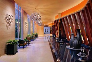 Un vestíbulo de hotel con un largo pasillo, ventanas amplias, plantas en macetas, un candelabro y jarrones decorativos.