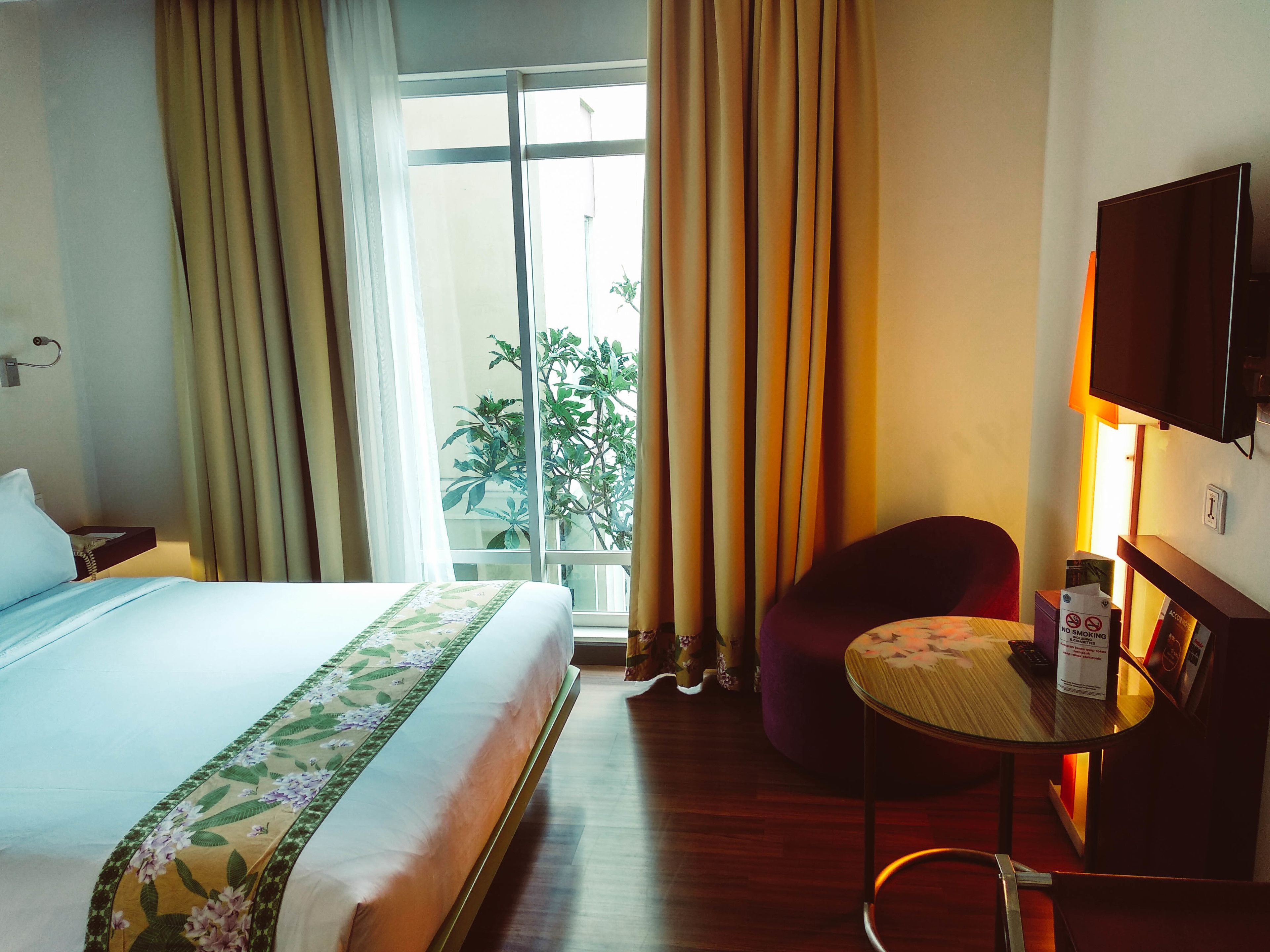 Habitación de hotel con cama, una silla, una mesita, televisor y ventana con cortinas.