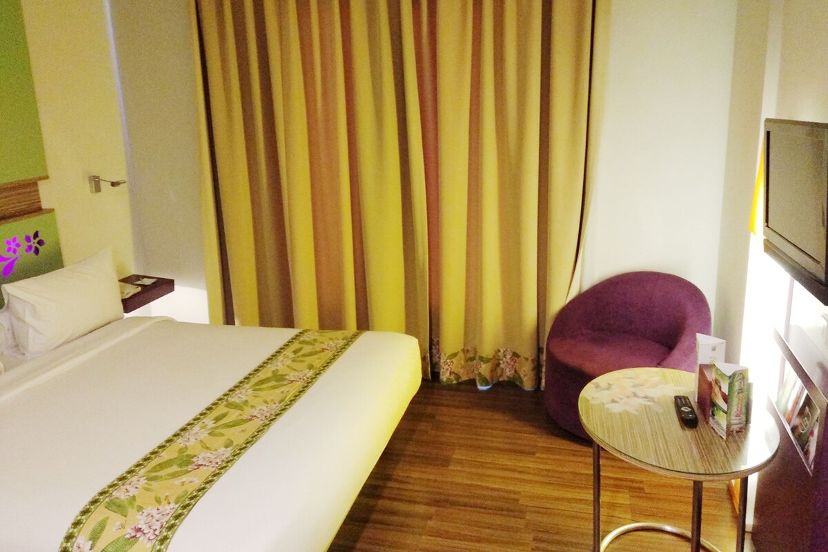 Una habitación de hotel con una cama, una mesita, una silla morada y un televisor en la pared.