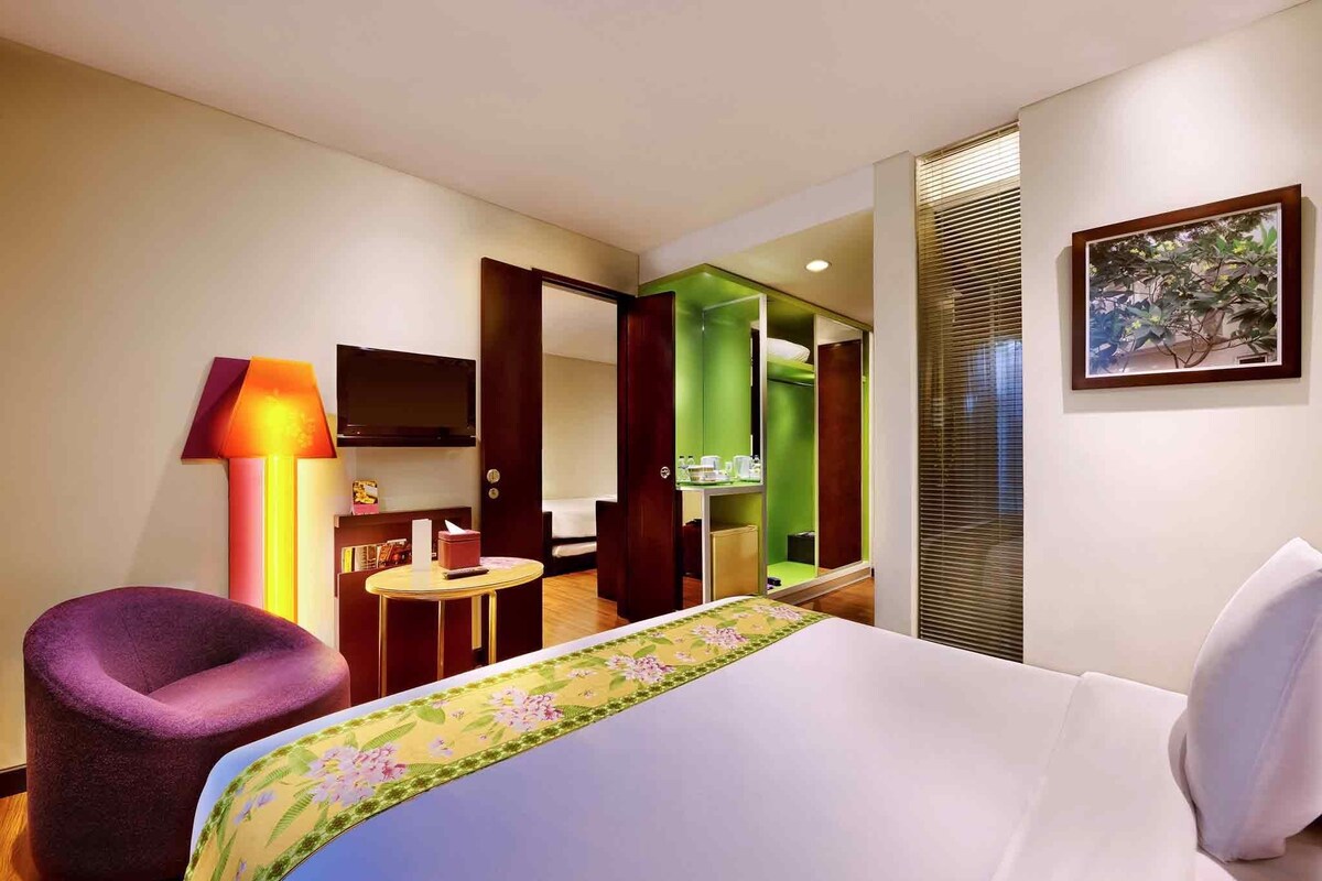 Habitación de hotel con cama, una silla morada, una mesita, televisión y un baño visible a través de una puerta abierta.