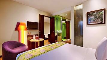 A hotel room with a bed, a purple chair, a small table, a TV, and a bathroom visible through an open door.