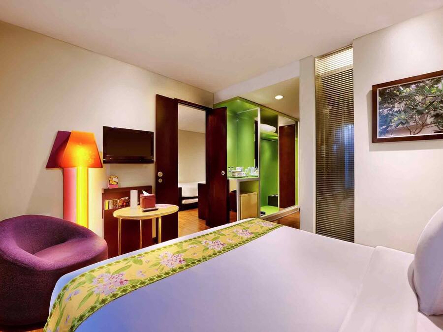 A hotel room with a bed, a purple chair, a small table, a TV, and a bathroom visible through an open door.