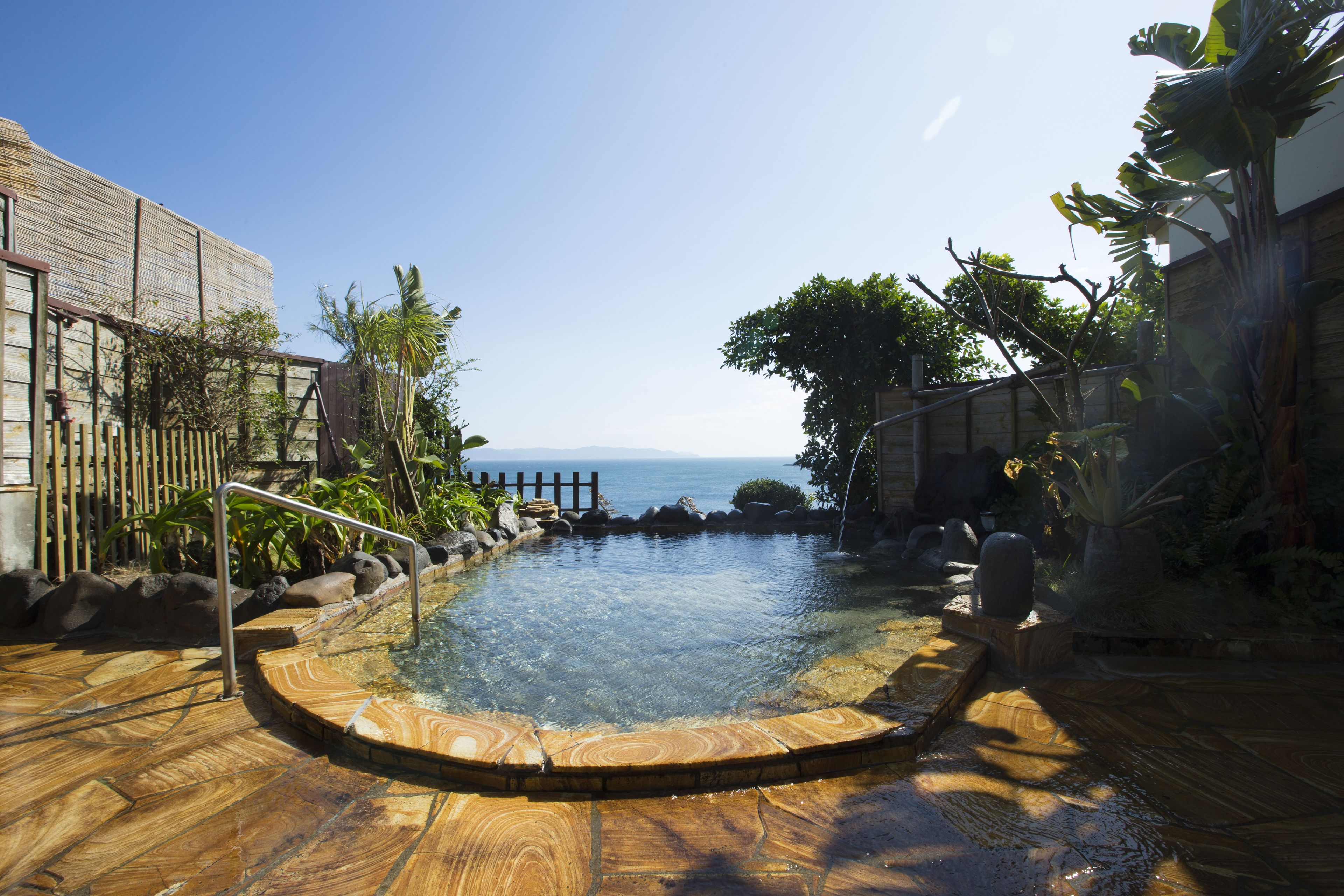 A pool with a wooden deck, surrounded by plants and a view of the sea.