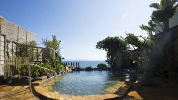 A pool with a wooden deck, surrounded by plants and a view of the sea.