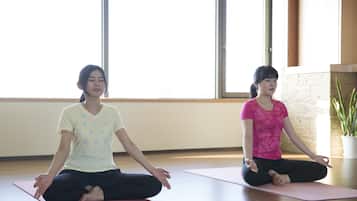 Two individuals seated on yoga mats in a room with large windows and a potted plant.
