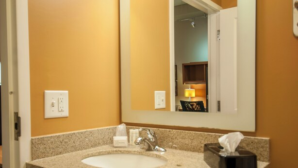 A hotel bathroom with a granite countertop sink, a mirror, and a towel dispenser. - TownePlace Suites by Marriott Baton Rouge Gonzales (Gonzales)
